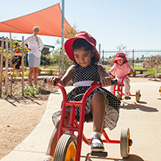 YMCA staff member monitors three children riding tricycles in an outdoor courtyard.